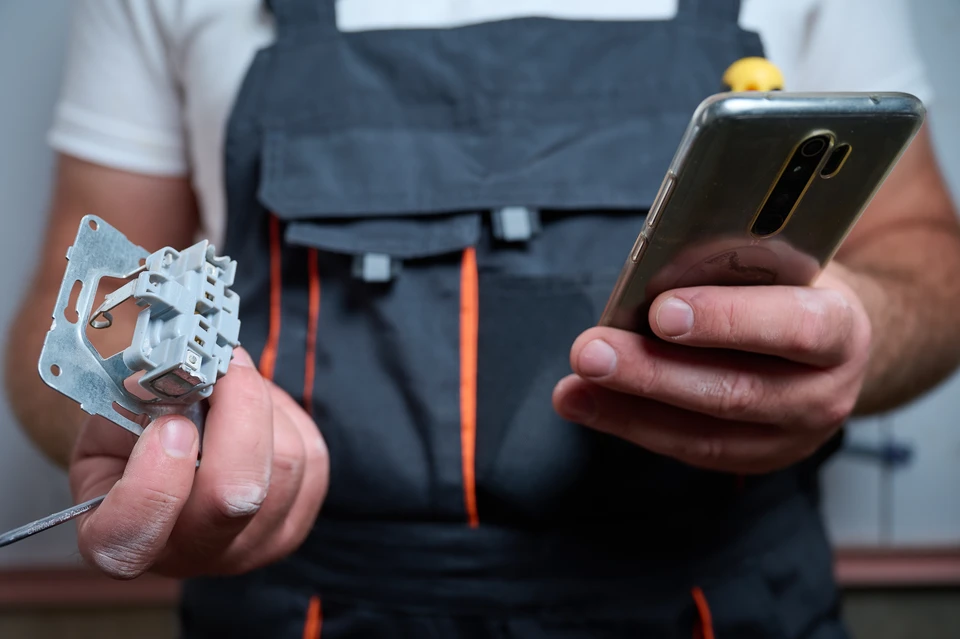 Photographie d'un électricien en uniforme professionnel travaillant à l'intérieur d'un logement résidentiel, effectuant une installation électrique visible (tirage de câbles, installation d'appareillage électrique mural, ou travail sur un tableau électrique). Le technicien est concentré sur son intervention. L'arrière-plan montre un intérieur résidentiel moderne et propre. La scène doit transmettre le professionnalisme, la compétence technique et l'engagement direct de l'artisan sur le chantier.